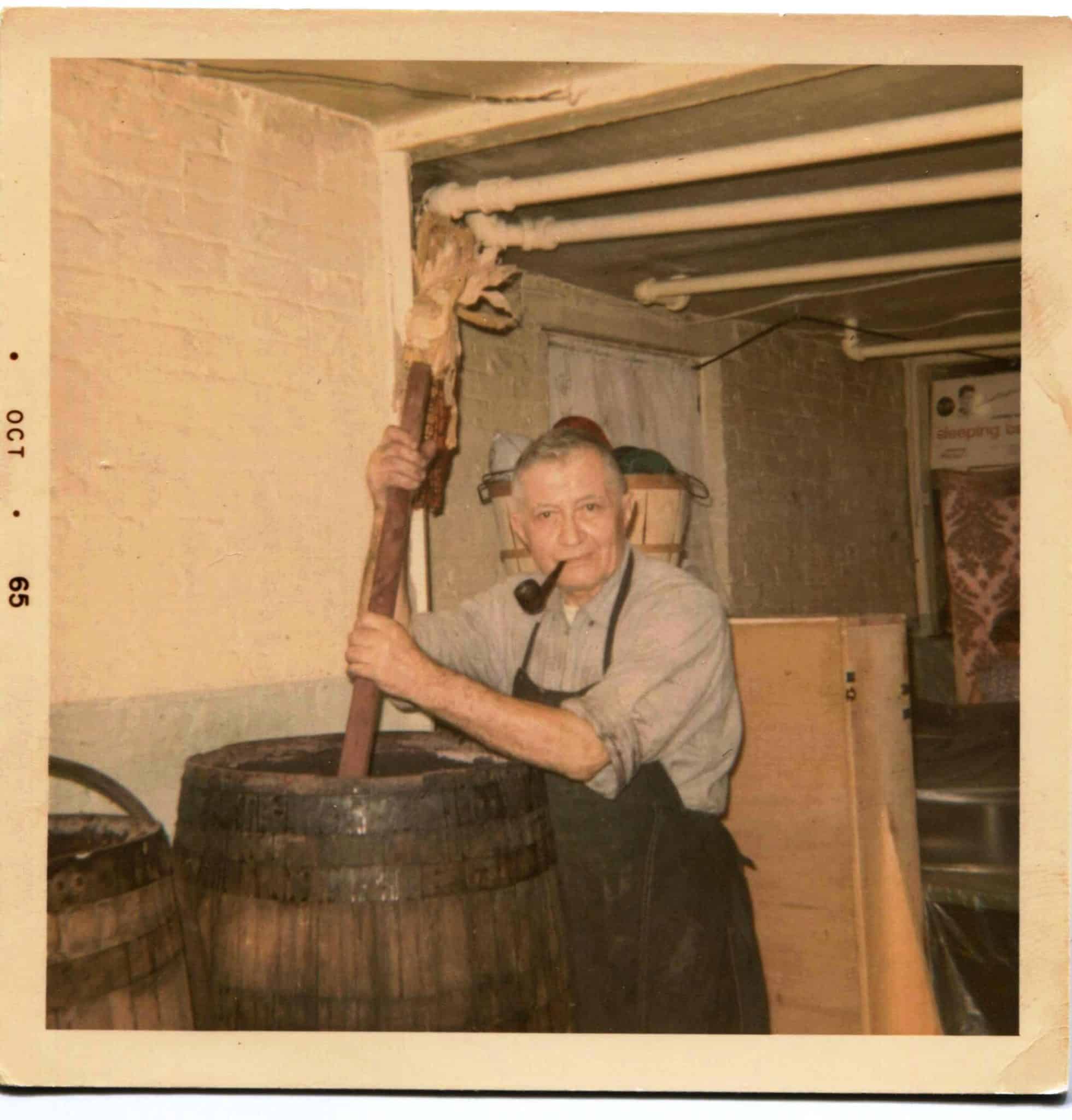 Image of Archimede Talevi, an older man with a pipe in his mouth holding a pole as he makes wine in a wooden barrel.
