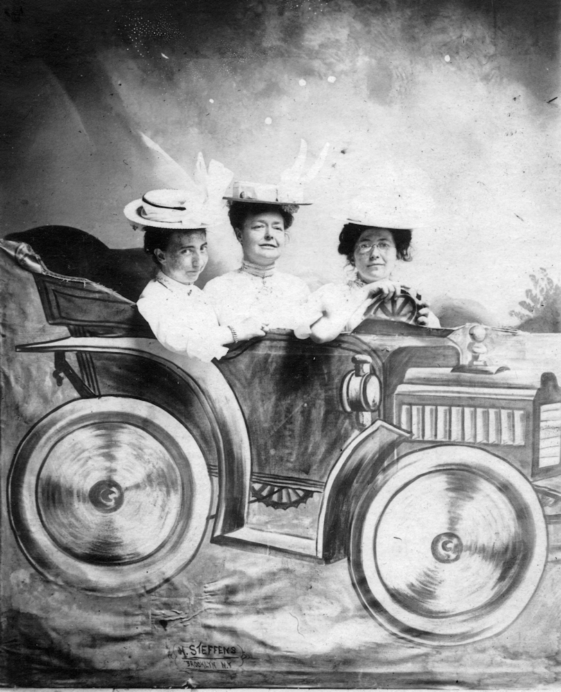 Three women in a black and white photo. They are in a cardboard car at a photo booth.