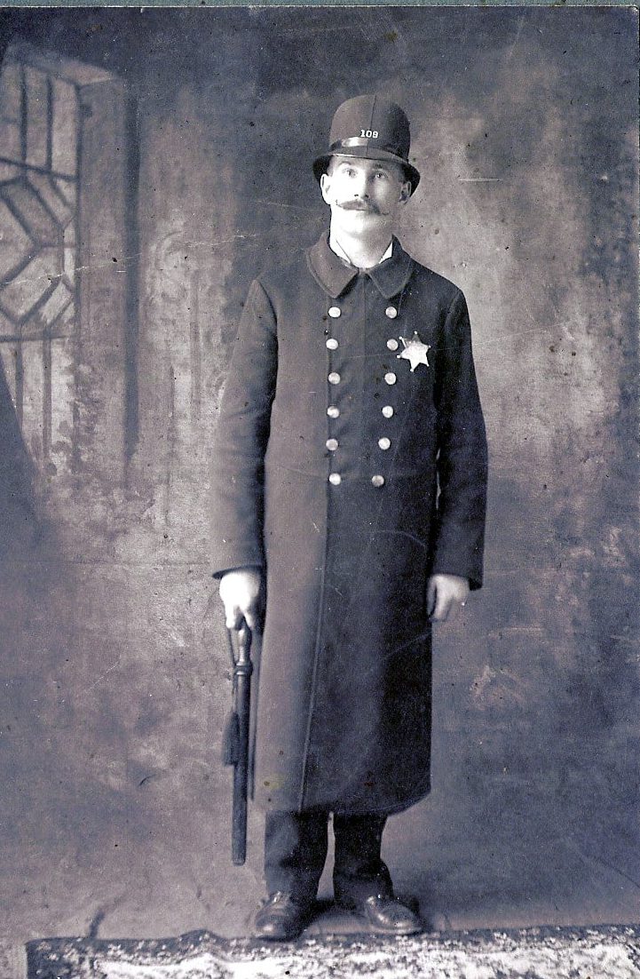 Man in a long police coat uniform, badge on his chest, and a weapon in his hand.